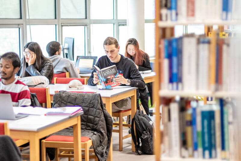 Photo de la médiathèque : à droite une étagère de livre, à gauche des étudiants qui travaillent ou lisent assis à des tables