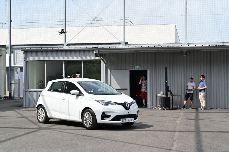 Two people are piloting a drone hovering above an autonomous car, which is itself being controlled by the drone