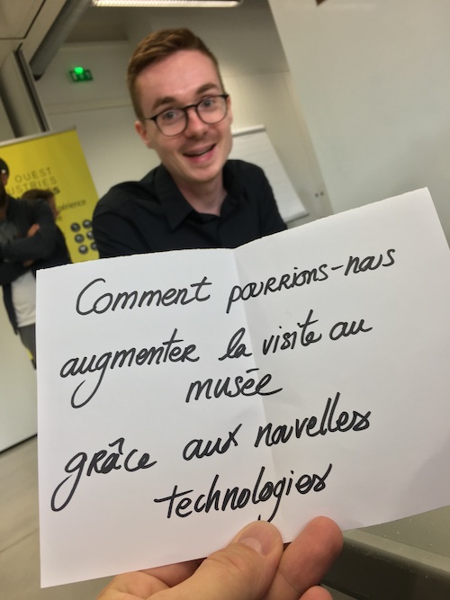 Une main tient un papier sur lequel il est écrit : Comment pourrions-nous augmenter la visite au musée grâce aux nouvelles technologies ?